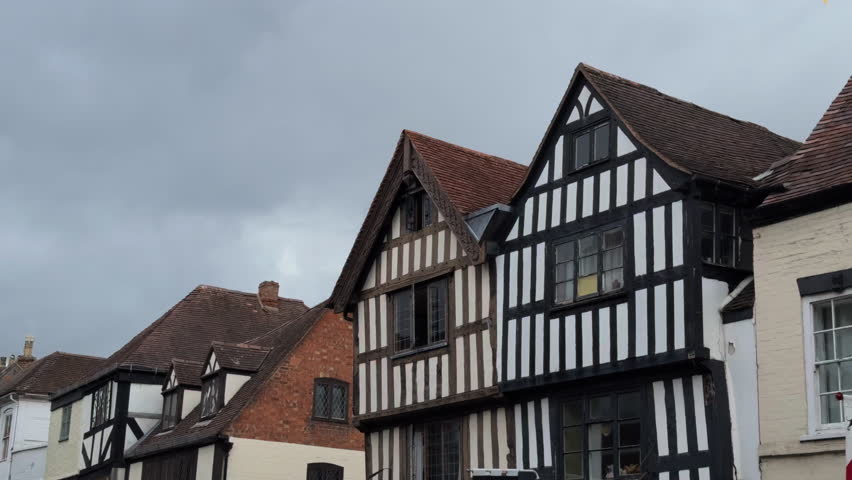 Static shot of a historic Tudor street with half-timbered facades, red tile roofs, and an overcast sky evoking stillness and age.