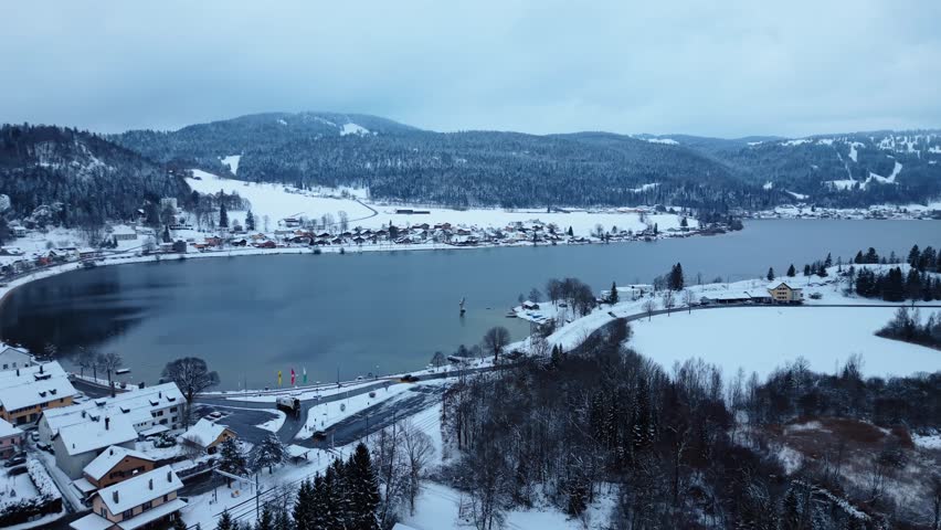 Aerial view of a serene Joux Lake surrounded by snow-dusted town and mountains under a cloudy sky, creating a tranquil winter scene, Le Lieu, Vaud, Switzerland.