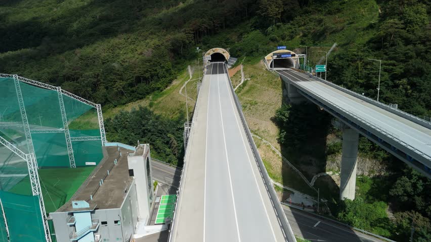Aerial view of a modern highway system with tunnels and an elevated bridge winding through lush green, forested mountains.