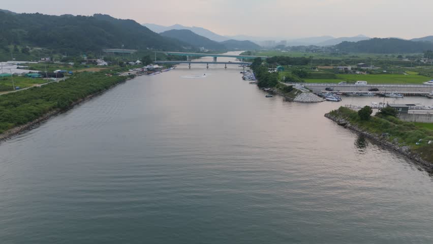 Aerial panorama of a wide river featuring a jet skier, multiple bridges, and diverse landscapes including green hills, greenhouses, a bustling marina, and distant city buildings, blending natural beau