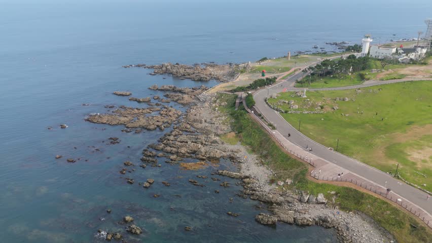 Aerial drone shot of a scenic coastal landscape featuring a lighthouse, winding road, and rocky shoreline.