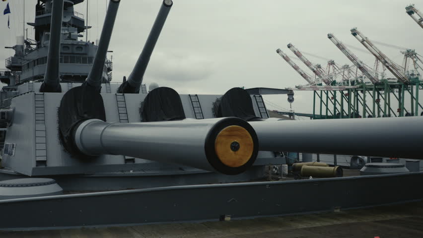 A dramatic tilt-up shot from the forward deck of a navy destroyer revealing multiple large naval guns. The camera moves upward from the deck to the imposing artillery turrets.