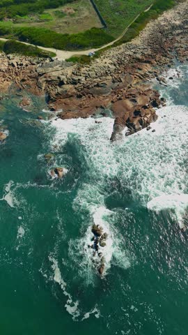 Aerial forward motion shot from above the ocean approaching the coastline. Waves breaking along a rugged rocky shore. A single camper van parked near the edge of the coast. Clear weather and soft natu