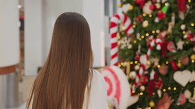 Back view of white woman walking towards festive Christmas tree with holiday decorations inside modern shopping mall, candy canes, stars, ornaments, and lights creating joyful atmosphere - Powered by Shutterstock - Get 15% off with code: PIKWIZARD15
