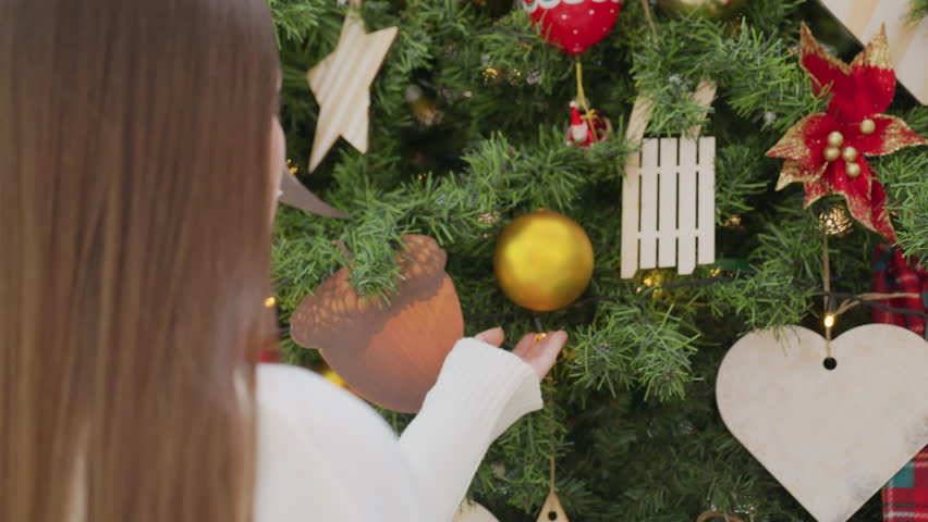 Hand view of lady in long-sleeve gently holding golden Christmas ball, observing it with soft bokeh lights in the background