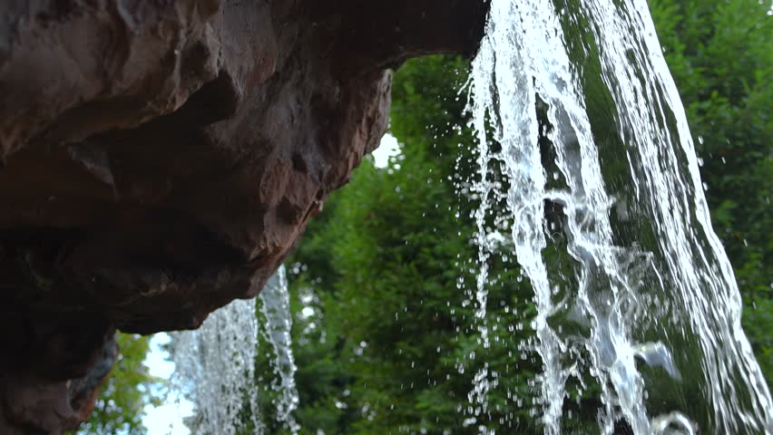 Garden Waterfall, Waterfall flowing from rocky cliff with cascading water drops, Close-up waterfall pouring over a rocky cliff with clear flowing water and lush green trees in the background