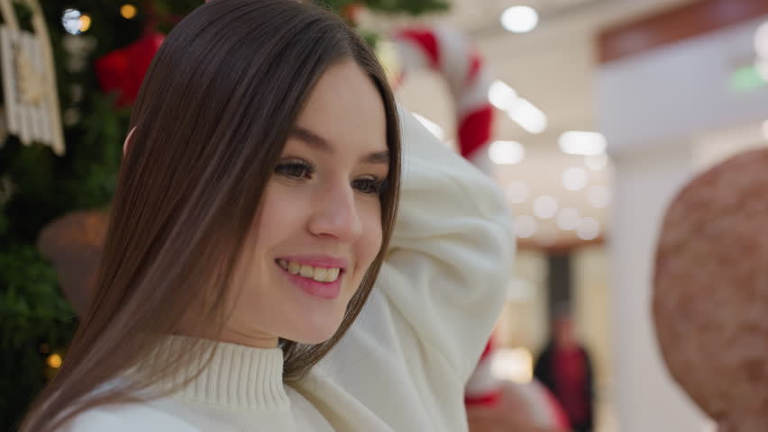Elegant lady in white sweater posing for a photo in front of Christmas tree in mall, with blurred background showing shoppers passing by