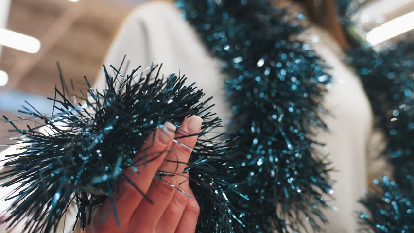 Close-up of person feeling texture of dark blue tinsel with blurred background, shimmering decor, and a passerby in a vibrant holiday shopping atmosphere, showcasing festive joy