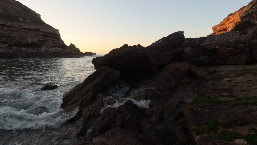 A low-angle shot of ocean waves washing over the rocky shoreline at Samarra Beach, with sunset light catching the cliffs and rough coastal textures