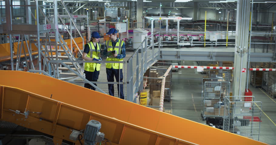 Two Warehouse Engineers in Monitor a Smart Parcel Conveyor, Using a Digital Tablet and Holographic Data Visualisations to Track Shipment, Optimise Distribution, Manage Logistics in Automated Hub.