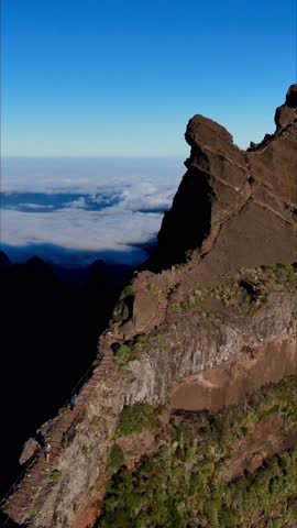 A vertical view of a narrow mountain ridge rising above a sea of clouds, with steep rocky cliffs, high-altitude trails, and vivid blue morning sky