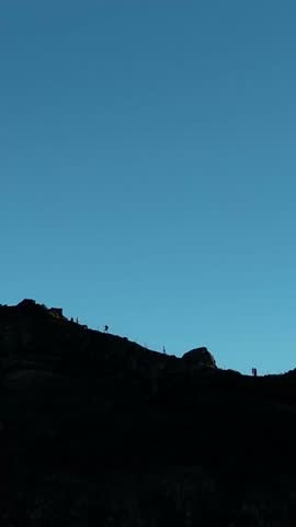 A vertical view of silhouetted hikers moving along a dark mountain ridge at dawn, contrasted against a clear pale sky with soft early-morning light
