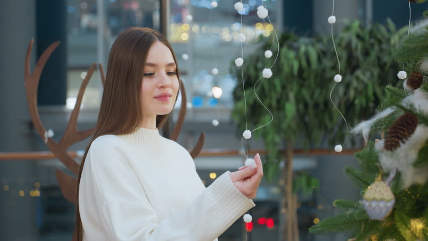 Woman in white sweater gently touches Christmas ornaments hanging on tree in mall, surrounded by festive lights and decorations