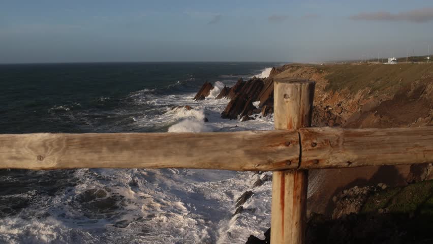 A coastal view with waves crashing into dark, jagged rock formations beneath a cliff along the coast of Sao Pedro of Moel, Leiria, Portugal - Powered by Shutterstock - Get 15% off with code: PIKWIZARD15