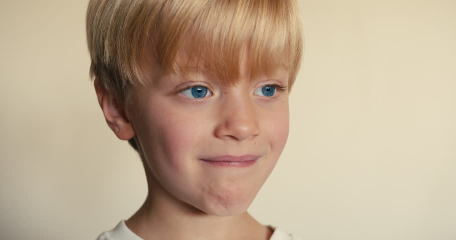 A close-up of a blond boy who smiles sincerely and touches his loose milk tooth with his pointer finger.