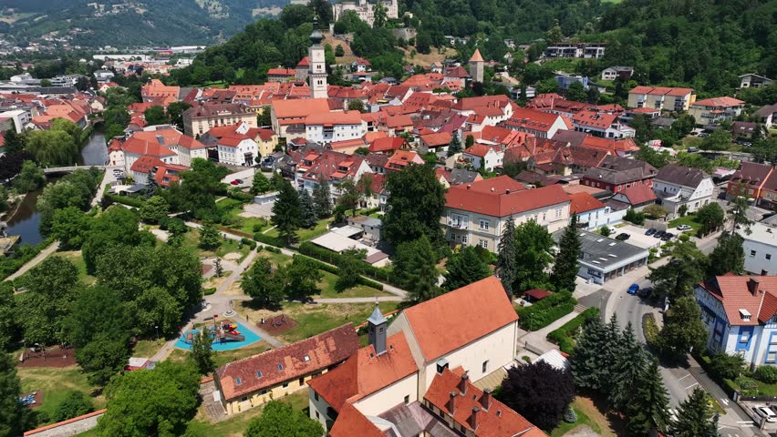 Aerial summer footage of Wolfsberg showing town center, church towers, green hills, layered valleys, warm light, smooth drone flight, atmospheric European small-town scenery