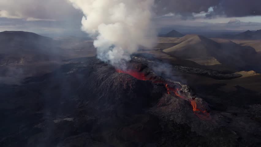 Wide aerial shot of active volcano releasing massive column of white gas and smoke into the sky. Red lava flows visible on the crater rim in vast barren landscape.