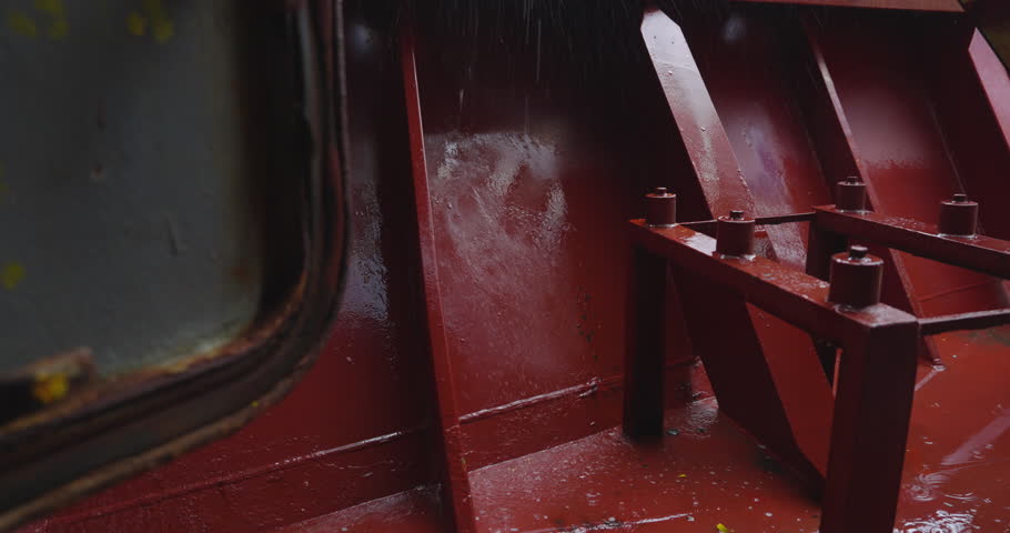 Scarlet metal beam glistens under fastly streaming water. Rust gleam merges with texture of rain salt and time. Flood on cargo vessel