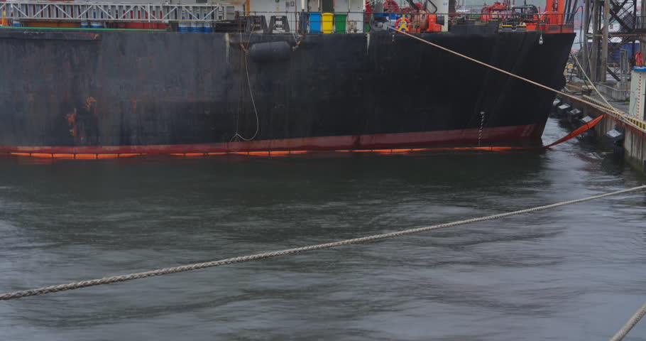 Giant freight vessel anchored by pier under gray sky. Water mirrors steel hull and distant harbor lights. Tanker stands at mooring line