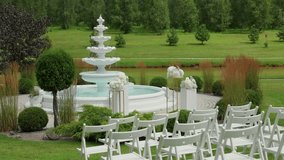 A peaceful garden wedding setup with white chairs arranged around a tiered fountain, surrounded by manicured shrubs and lush green scenery - Powered by Shutterstock - Get 15% off with code: PIKWIZARD15