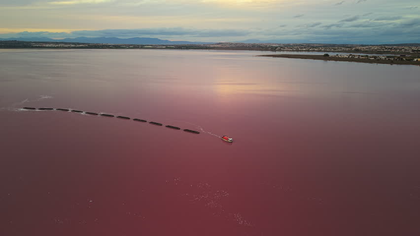 Aerial view of a red boat towing black barges across a pink salt lake at sunset, with mountains and a distant shoreline under a cloudy sky. High quality 4k footage