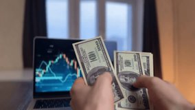 A man's hand holds several dollar bills, where dollar bills are prominently displayed before a laptop screen. The laptop screen shows financial graphs near the dollar bills. - Powered by Shutterstock - Get 15% off with code: PIKWIZARD15