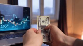 A man's hands hold dollar bills while counting dollar bills in front of a laptop screen. The laptop screen displays financial graphs next to the dollar bills held by the man. - Powered by Shutterstock - Get 15% off with code: PIKWIZARD15