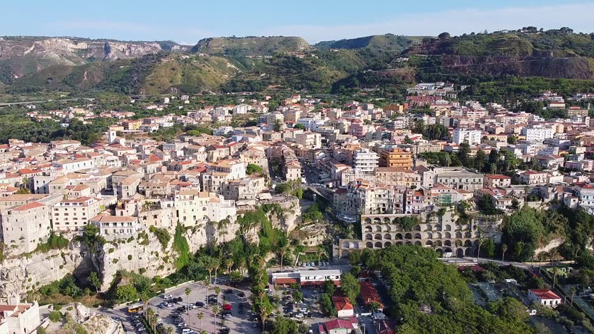An aerial wide shot reveals Tropea, Italy, with its dense urban center perched atop dramatic seaside cliffs. Lush green hills and mountains form a scenic backdrop.
