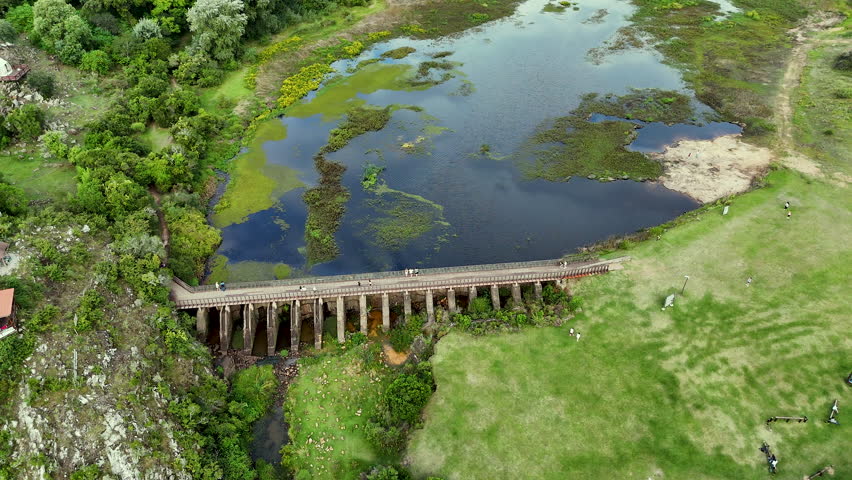 Aerial view of an old bridge and lagoon in Villa Serrana, Uruguay. Drone footage shows green landscape, calm water and people walking over the historic structure.
