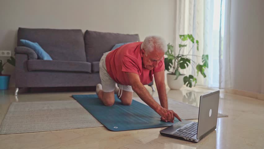 Senior man in sportswear using laptop and doing plank exercise on yoga mat at home