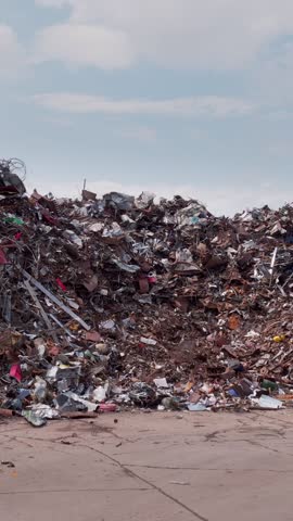 Vertical video. Large mound of scrap metal stands in recycling yard by cracked concrete under sky. Twisted beams wires and rusted sheets form mixed debris while site awaits sorting processing.