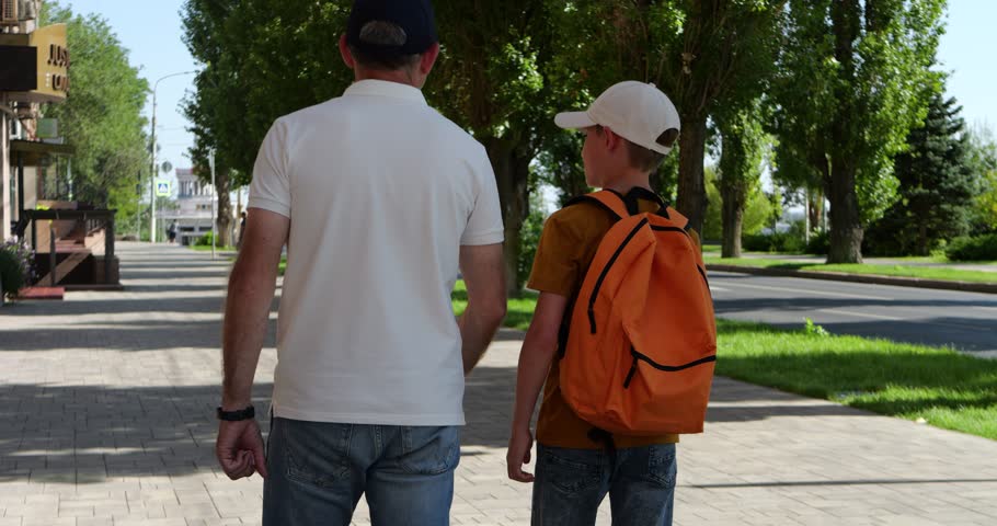 Father and son walk outdoors on sunny summer street. Dad and child talk and enjoy calm family moment. warm family bonding. Green trees create peaceful mood. Family theme feels positive and natural.