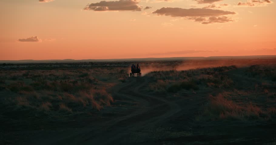 Distant cinematic shot of riders with a donkey cart at sunset, their silhouette framed by orange backlit dust rising from the desert road in Namibia.