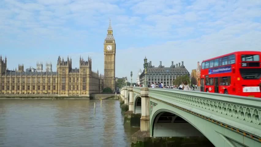 View captures Big Ben standing tall against a blue sky, buses crossing Westminster Bridge, people walking along the riverbank, and historic structures lining the waterfront.