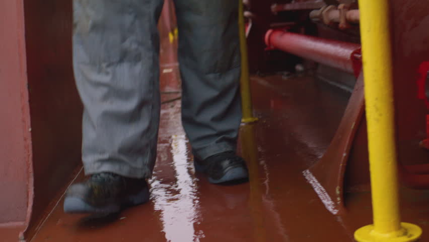 Man in heavy boots walks steadily across wet deck while reflections shimmer under gray sky. Worker steps through puddles on metallic surface