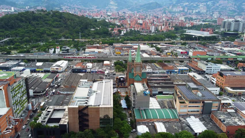 Downtown Medellin scene with metro train passing through dense urban buildings, active streets, mixed architecture and bright daylight, capturing a dynamic central corridor in a wide city panorama tod