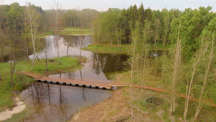 Aerial view of a peaceful natural landscape in rural Lithuania featuring a small pond or lake, lush green trees, and a simple wooden bridge connecting the banks.
