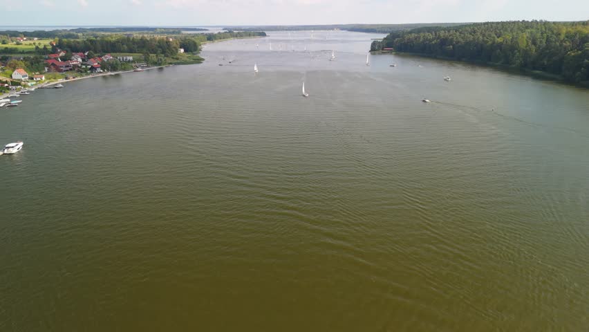 A serene view of Mikolajki, Masuria featuring a wide lake surrounded by lush greenery and sailing boats gliding across the water under a clear blue sky during a sunny day.