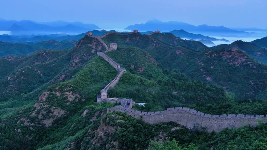 Great Wall stretches across lush green mountains under soft morning light, with mist rising in the distance creating a serene and picturesque atmosphere.