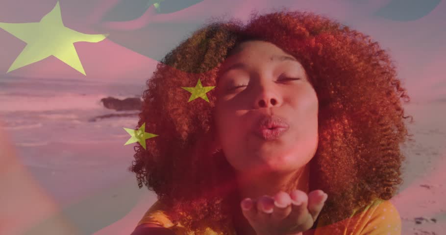 Woman blowing kiss filming social media at beach, causing stars to merge into stripes over face. Selfie, promotional, vibrant, branding, shoreline, outdoor, flag