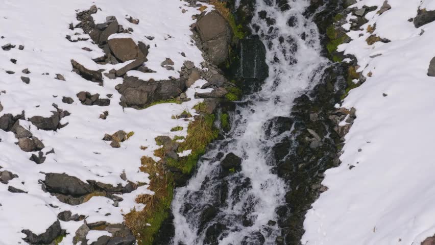 A rapid mountain stream flows over rocks and snow covered slopes in the North Caucasus region of Russia.