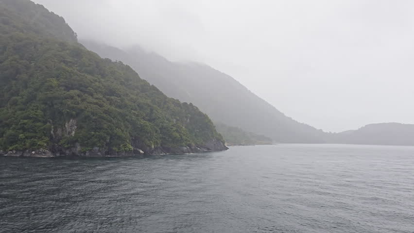 Milford Sound fiord with steep, lush mountains rising from calm water under misty, overcast skies, moody, serene scene of New Zealand
