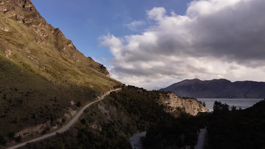 Aerial drone view of a winding gravel road on a steep hillside overlooking Lake Hawea. Dramatic mountain landscape and cloudy sky in the South Island of New Zealand