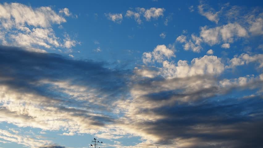 Yellow orange clouds drift across a bright blue sky as sunlight breaks through, creating a beautiful display of light and shadow. Slow motion, real time. TV antenna with birds. Evening sunset.