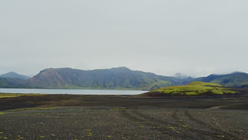 An aerial or wide angle shot of a serene lake surrounded by rugged, colorful mountains and a dark volcanic ash field. The scene captures the unique and dramatic natural beauty of the Icelandic Highlan