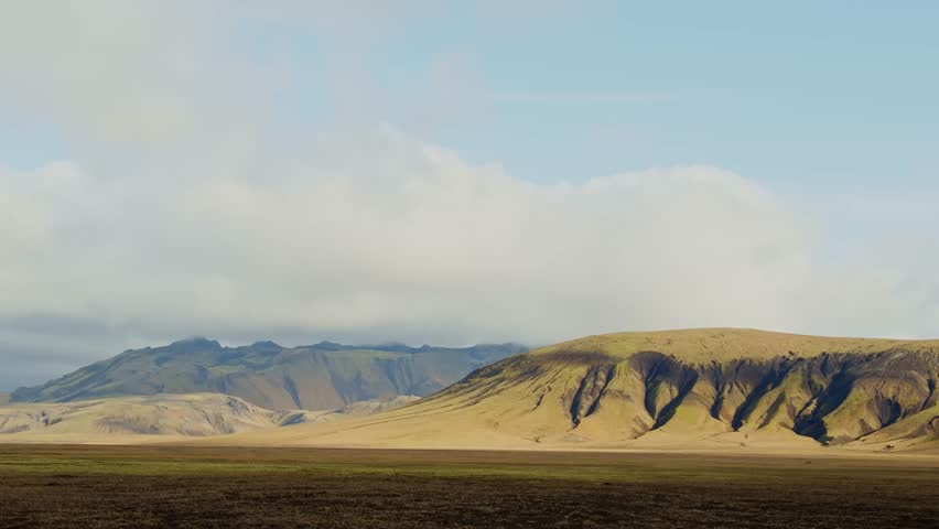 A wide-angle view of a classic Icelandic landscape with an open field of brown and green earth, rolling hills, and distant mountains beneath a dramatic, cloudy sky.