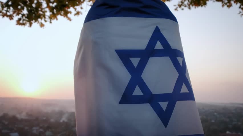 A woman stands with the flag of Israel against a clear sky at sunset, capturing national pride, symbolic gesture, and scenic evening light.