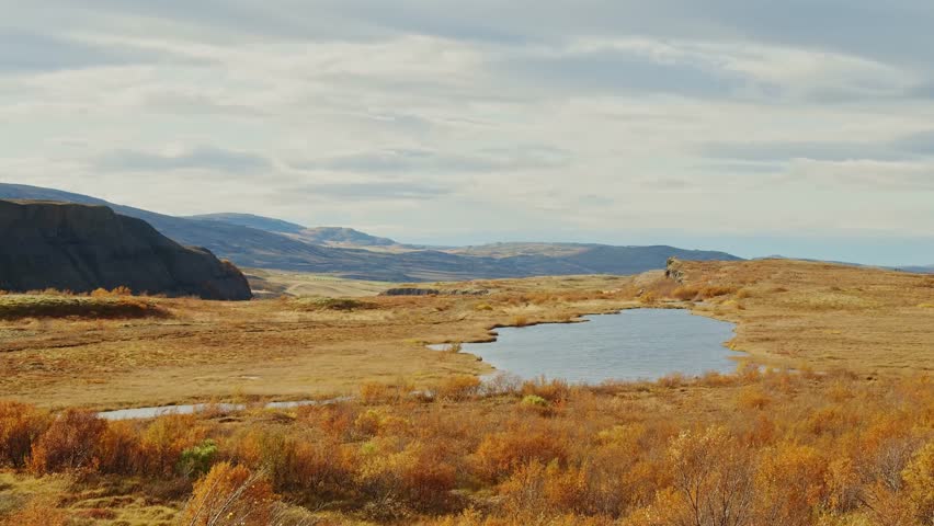 A wide-angle view of a serene Icelandic autumn landscape with a calm lake reflecting cloudy skies, surrounded by fields of golden and reddish-brown vegetation and distant hills.