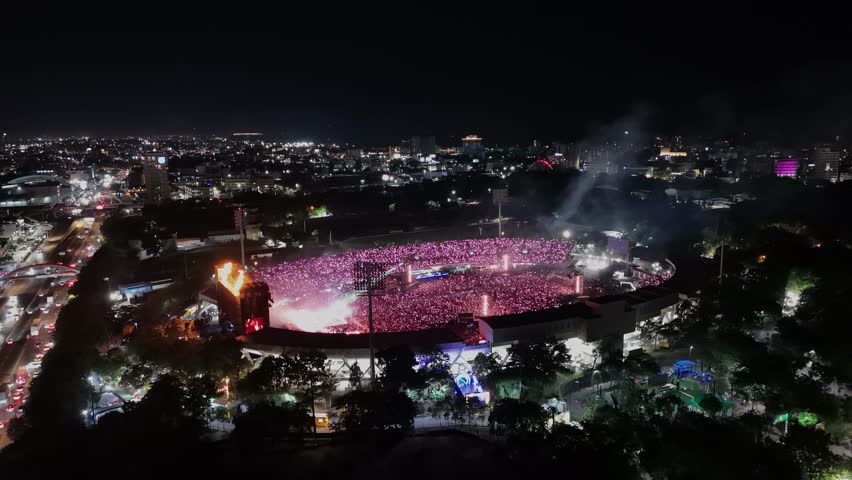 Aerial approaching shot showing crowd of people celebrating bad bunny festival show in open air stadium at night. Santo Domingo, Dominican Republic. Music show at night with flashing illuminated light
