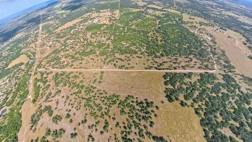 Aerial view from a small yellow airplane flying over dry countryside. The footage captures rugged terrain, scattered trees, dirt roads, and wide open farmland on a sunny day.